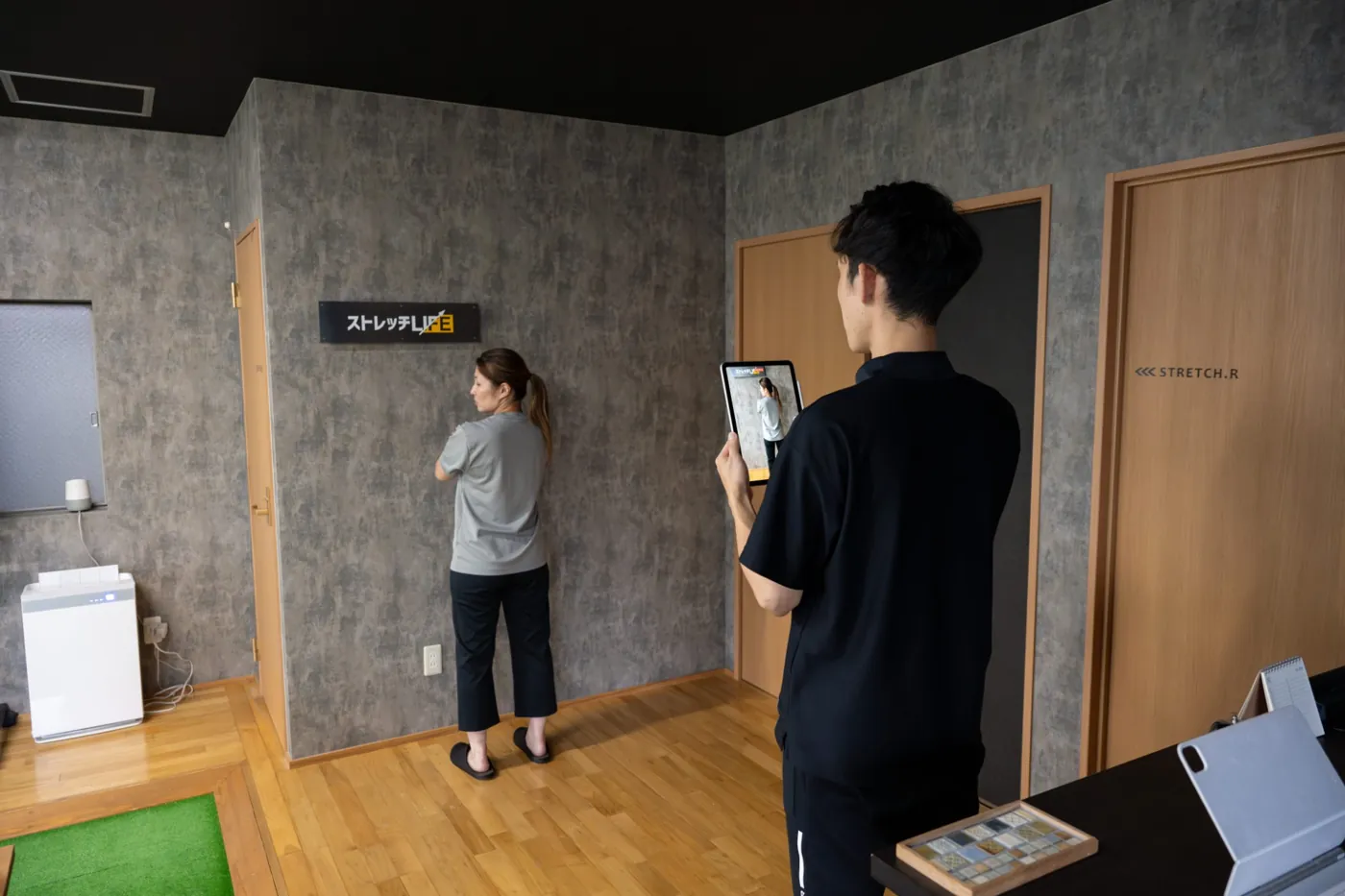 壁の前に立つお客様の姿勢を、トレーナーがタブレットで撮影して分析している様子