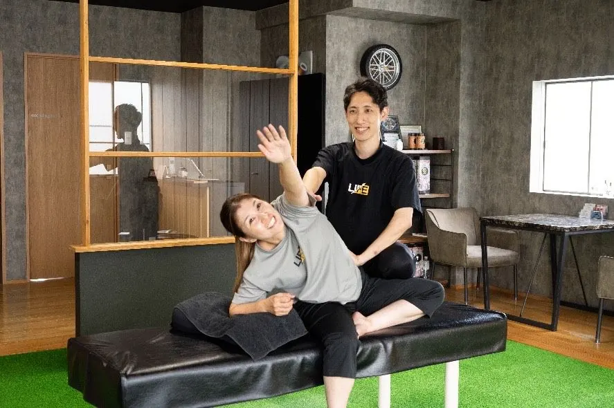 トレーナーに支えられて体を横に伸ばし、笑顔を見せる女性